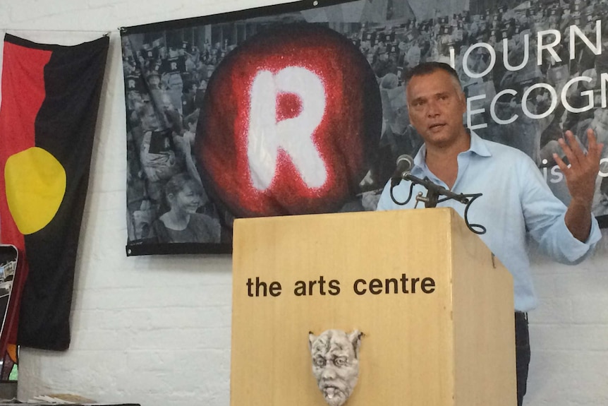 Stan Grant standing at lectern with Aboriginal flag and the word "Recognise" behind him