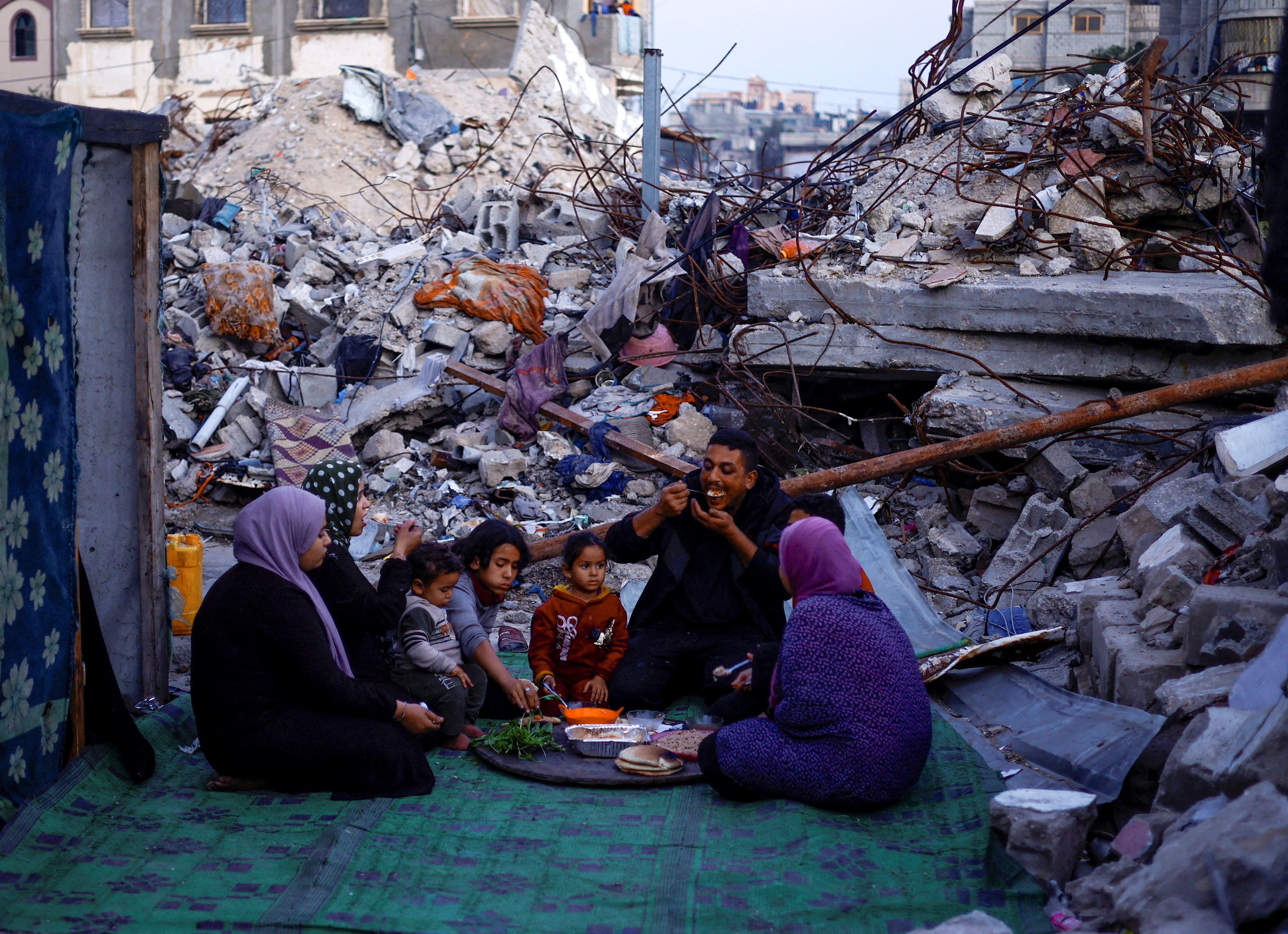 Family of eight sits in a semi-circle eating from a platter of food, surrounded by rubble.