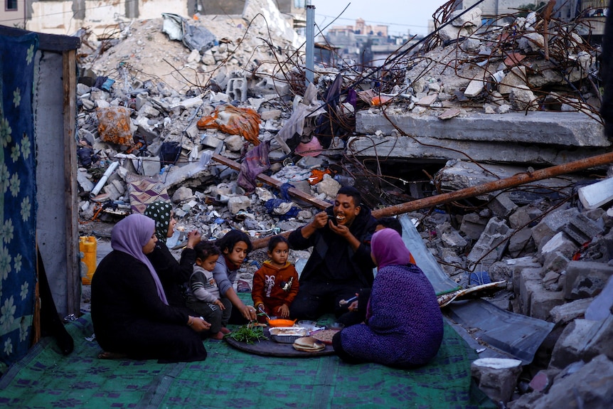 Family of eight sits in a semi-circle eating from a platter of food, surrounded by rubble.