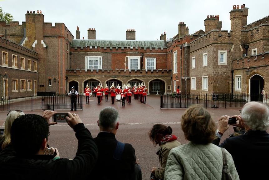 Palace exterior, guards in red with gold hats, royal fans taking photos