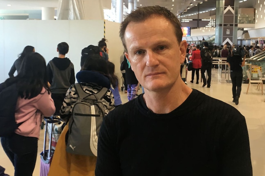 Dave Parry looks at the camera, with the airport in the background filled with passengers.