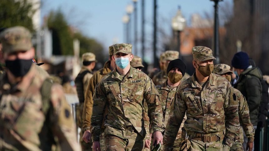 A group of soldiers wearing army uniforms and hats and masks walk together on a Washington street.