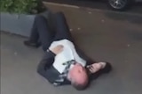 Grainy footage shows a man in a suit lying on the ground, one hand to his ear holding a phone.