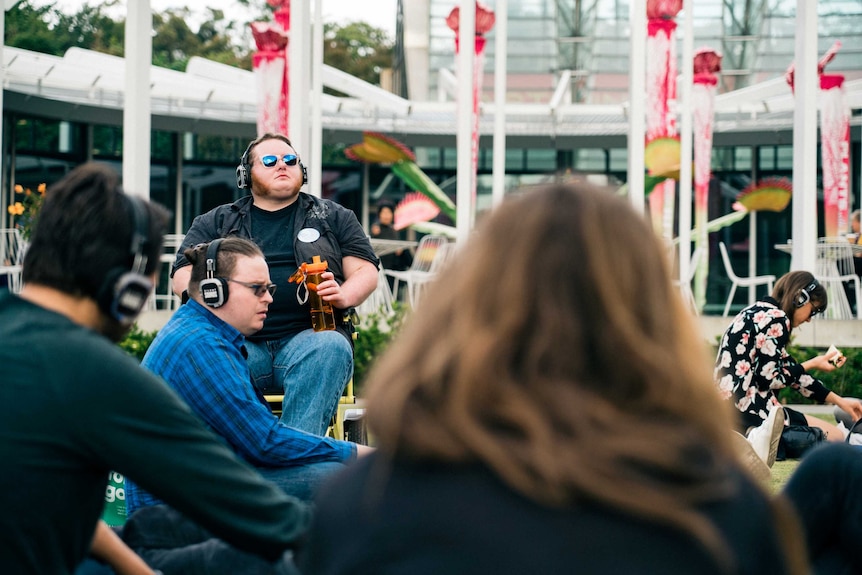 A man with sunglasses and drink bottle in a wheelchair with big headphones on, plus more people with headphones on in front