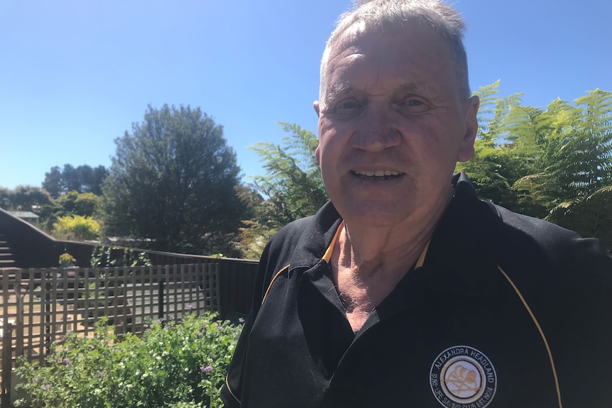 An older man wearing a black polo shirt looks at the camera.