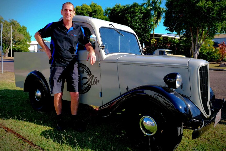 A man in work shorts and shirt leans against a classic historic ute parked in suburban street.