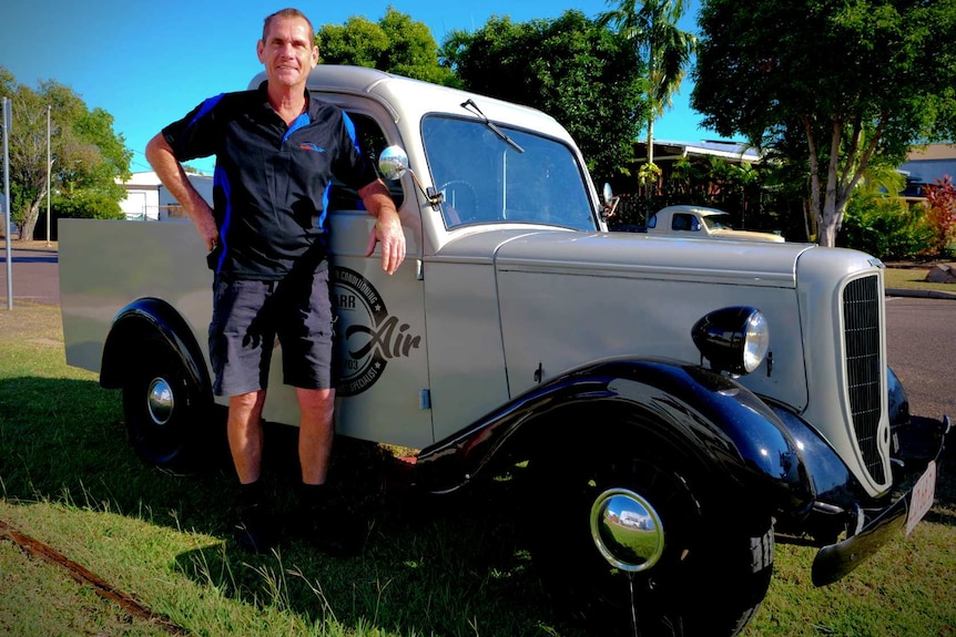 A man in work shorts and shirt leans against a classic historic ute parked in suburban street.