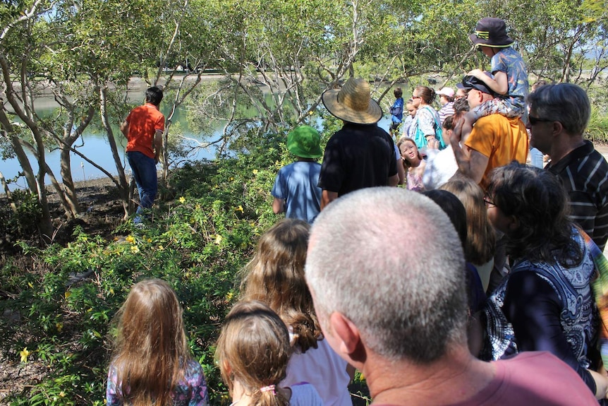 A circle of families stand watching a man who is walking through a mangrove toward an electronic sensor.
