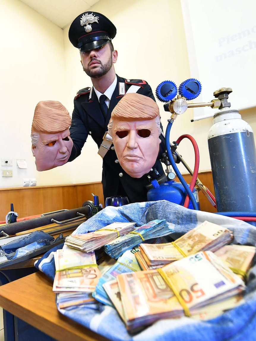 A uniformed police officer holds two Donald Trump masks used during bank robberies, behind a bag of euros.