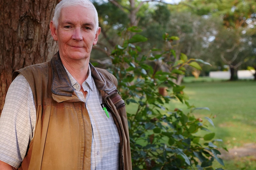 A man wearing a parker jacket stands in front of a tree and smaller bush in a park 