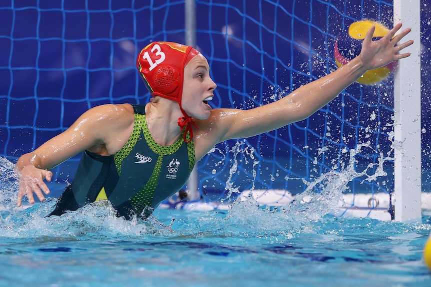 An Australian female water polo goal keeper stretches out with her left arm to try to stop a ball.