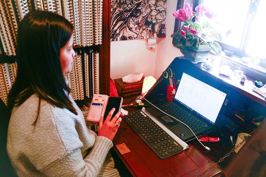 Ria using her phone as she sits at her desk, decorated by a pot of pink flowers and other ornaments.