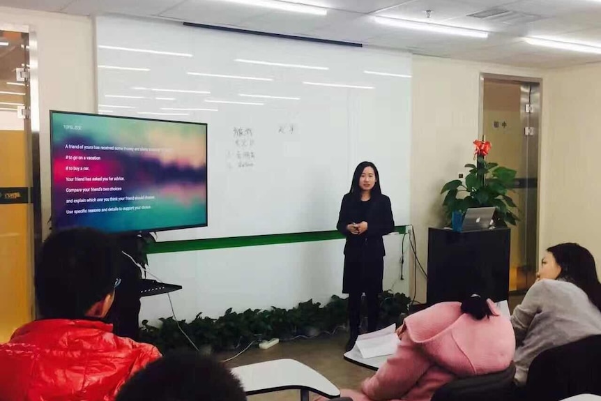 A young woman stands in front of a whiteboard and tv screen addressing an audience.