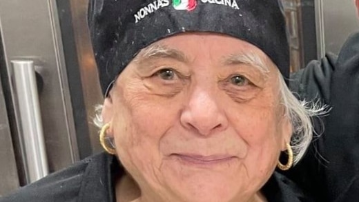 A close up of an older woman wearing a black chef's hat and apron reading 'Nonna's Cucina'