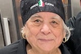 A close up of an older woman wearing a black chef's hat and apron reading 'Nonna's Cucina'