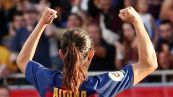 A footballer with her back to camera raises her arms in triumph as the crowd celebrates.