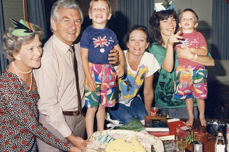 An old film photo of Bob Hawke with his children and grand-children, cutting a cake during a birthday party.