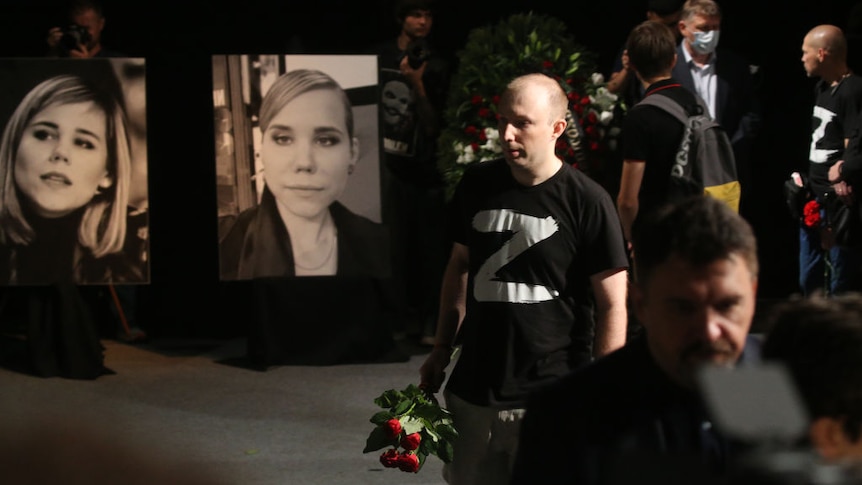 A man wearing a t-shirt with letter 'Z', a symbol of support of Russian invasion holds flowers. Images of Daria Dugina behind 