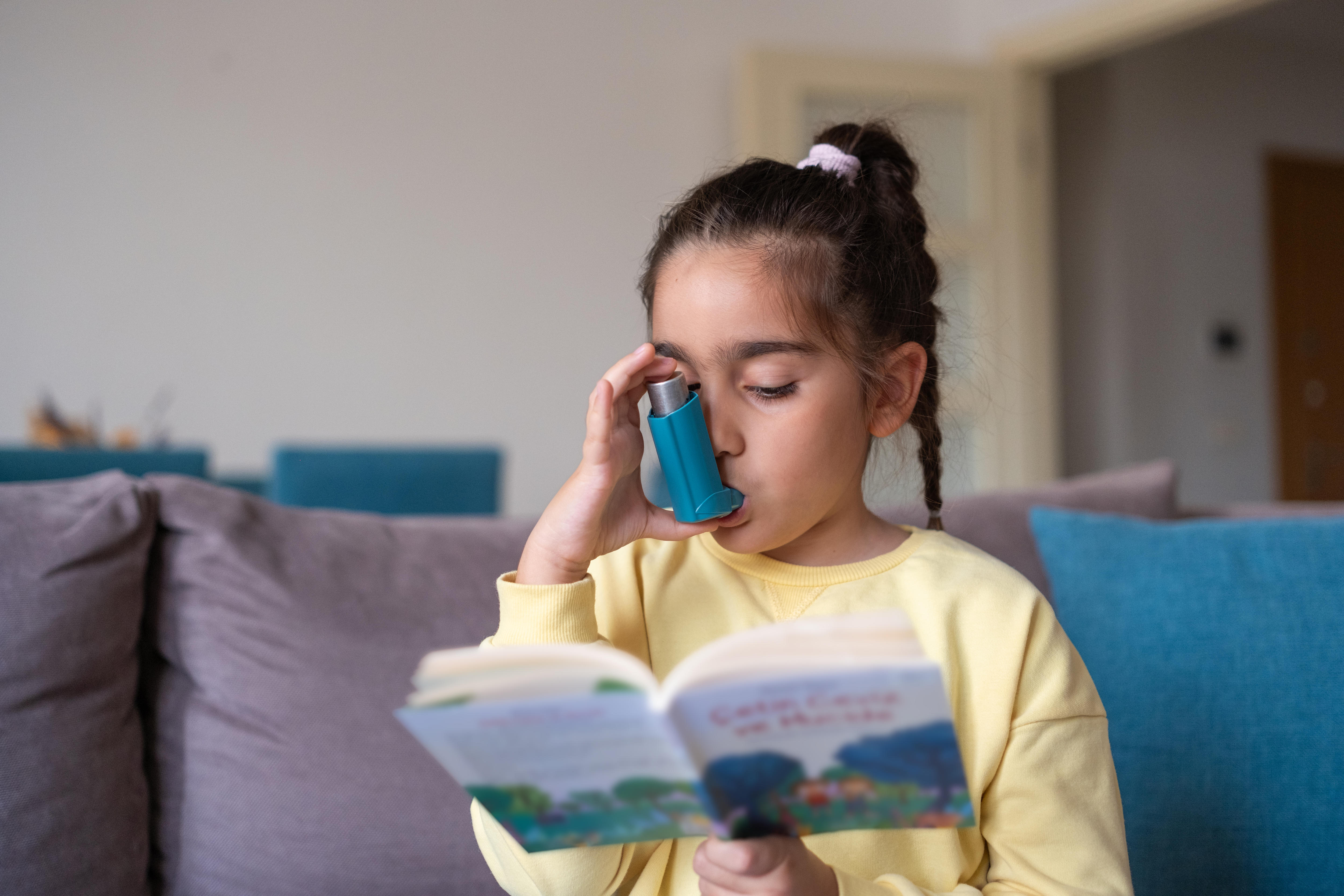 A little girl with dark hair is reading a book while she uses her asthma inhaler