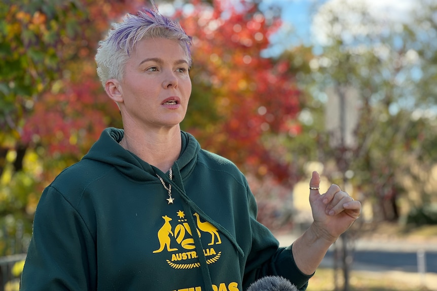 A woman with short hair wearing a Matildas hoodie speaks in front of a microphone