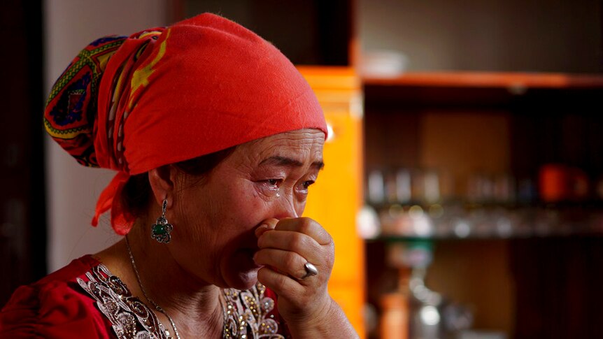A woman wearing a red headwrap and top holds her hand to her face as she wipes away tears