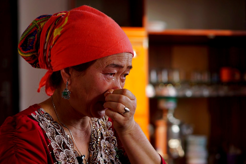 A woman wearing a red headwrap and top holds her hand to her face as she wipes away tears