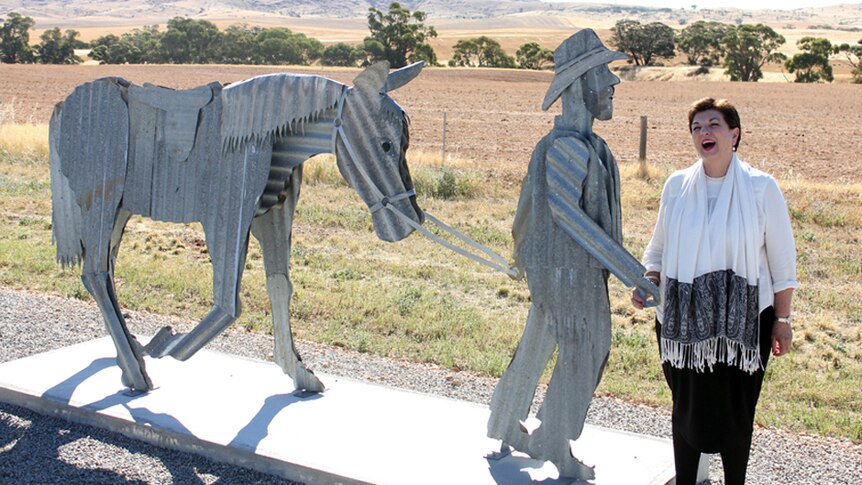 A woman laughing standing next to a corrugated iron statue of a man leading a horse.