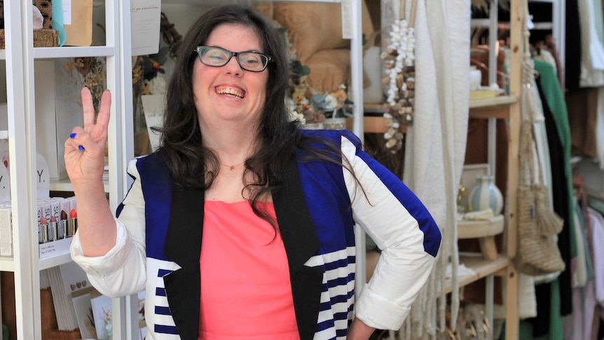 Simone wears a hot pink top, blue and white blazer and black glasses, posing with a peace sign in front of retail shop shelves. 