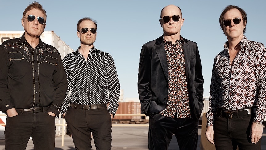 Four members of Hoodoo Gurus wearing sunglasses and button up shirts, standing outside