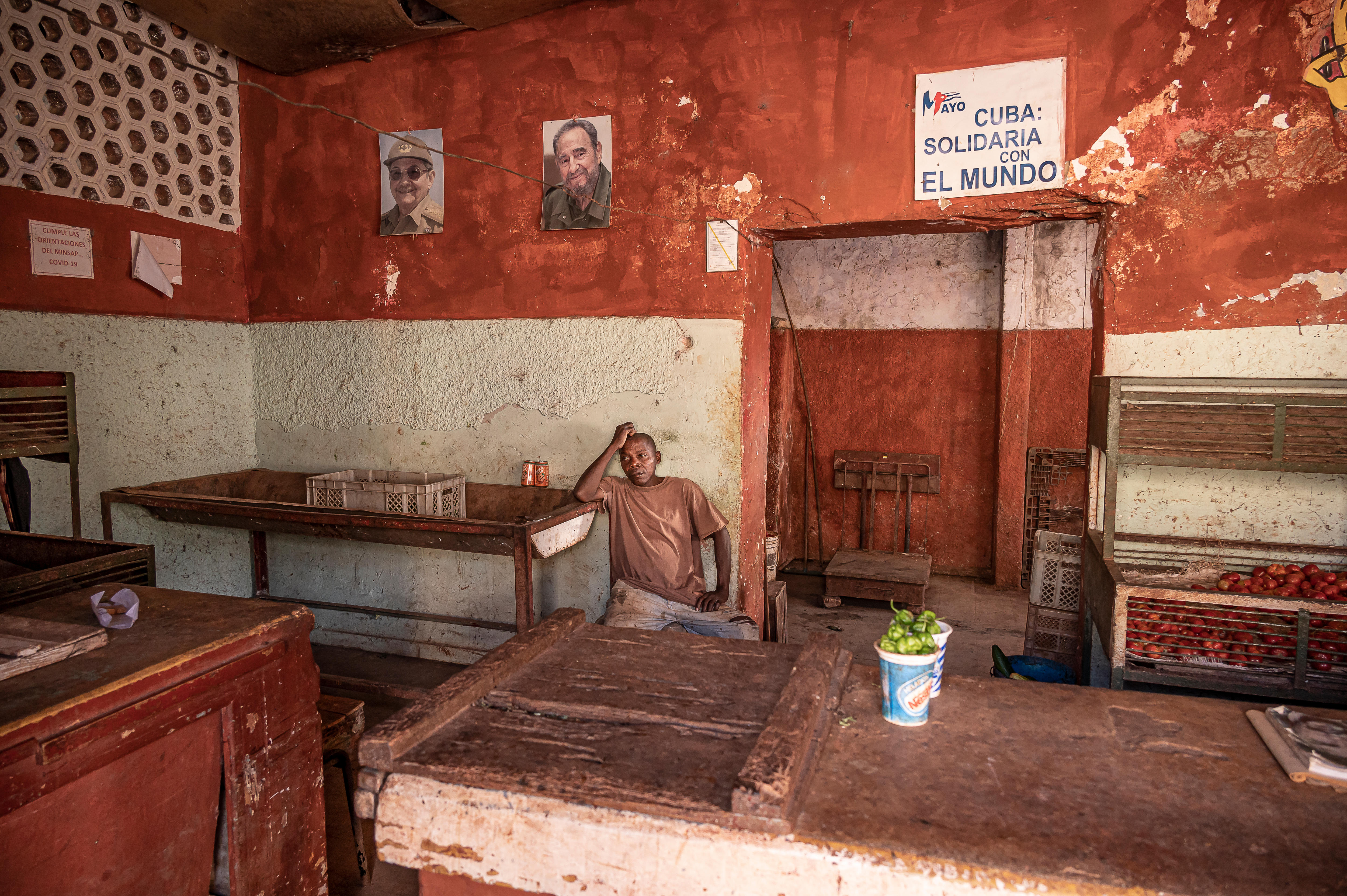 A Cuban shopkeeper rests against the red and white back wall of a greengrocer. The shelves are bare.