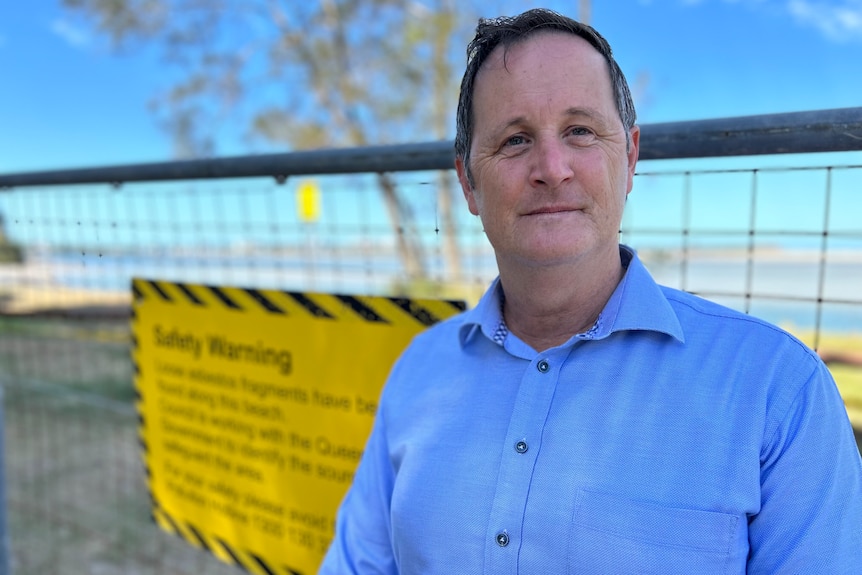 A man with short, dark hair, wearing a business shirt, stands in front of a fence with a warning sign attached to it.