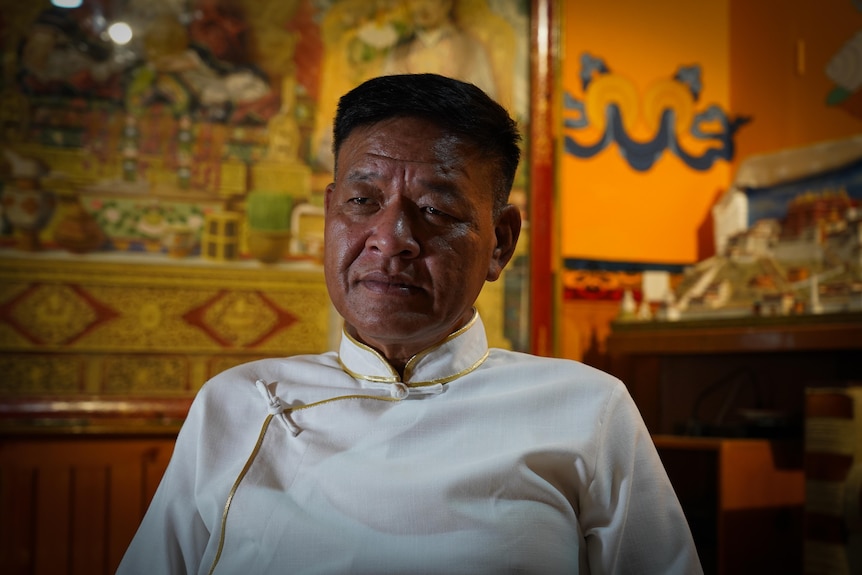 A man in white band-collared shirt trimmed with gold sits in an ornate room in front of a model palace