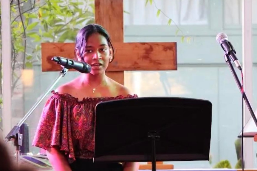 Satara speaking at church, on the stage with a wooden cross behind her.