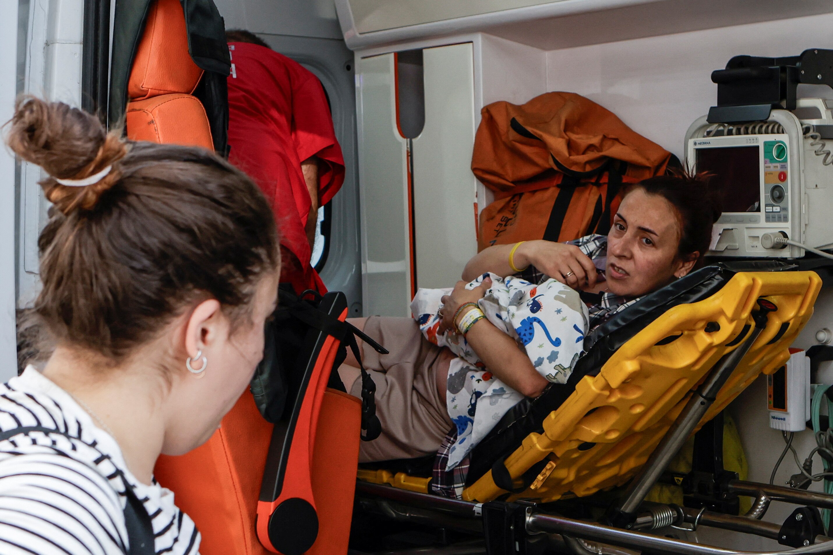 A woman holds a baby while laying in the back of an ambulance while speaking to another woman