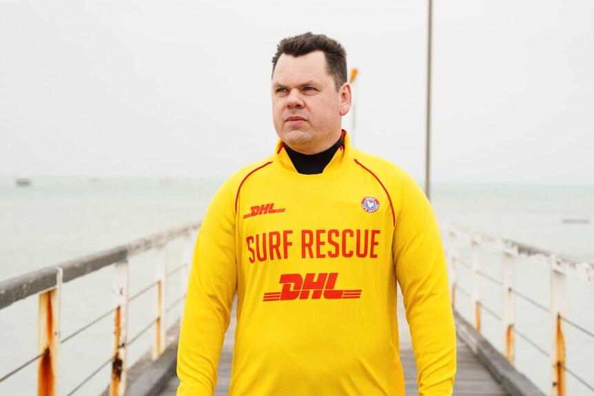 A man in a yellow 'surf rescue' swim shirt stands on an old jetty looking over the water.