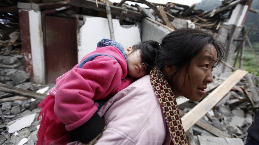 A survivor carries her sleeping daughter as she walks past debris at a village up on the mountainside.