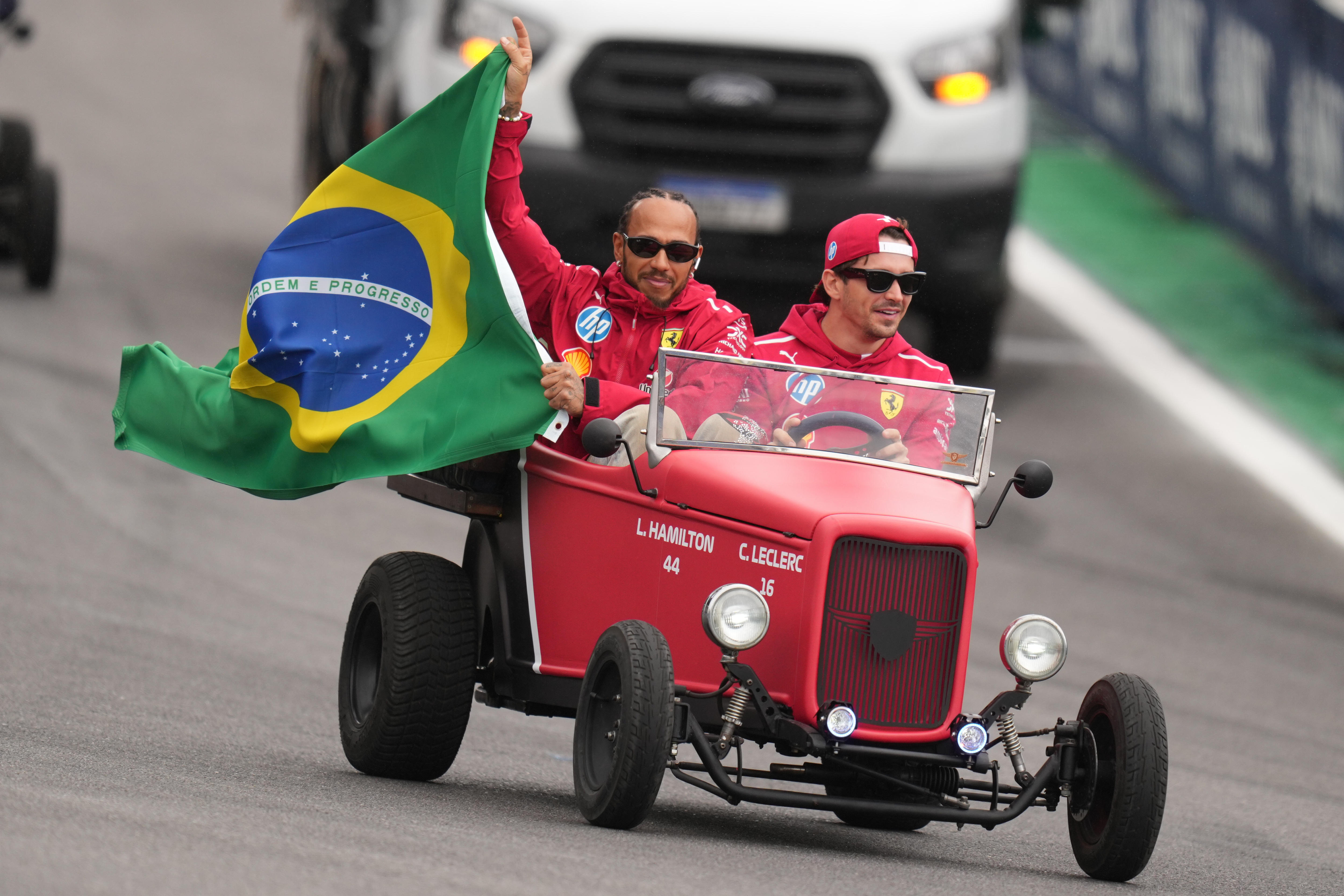 Lewis Hamilton e Charles LeClerc seguram uma bandeira do Brasil enquanto dirigem um carro antigo