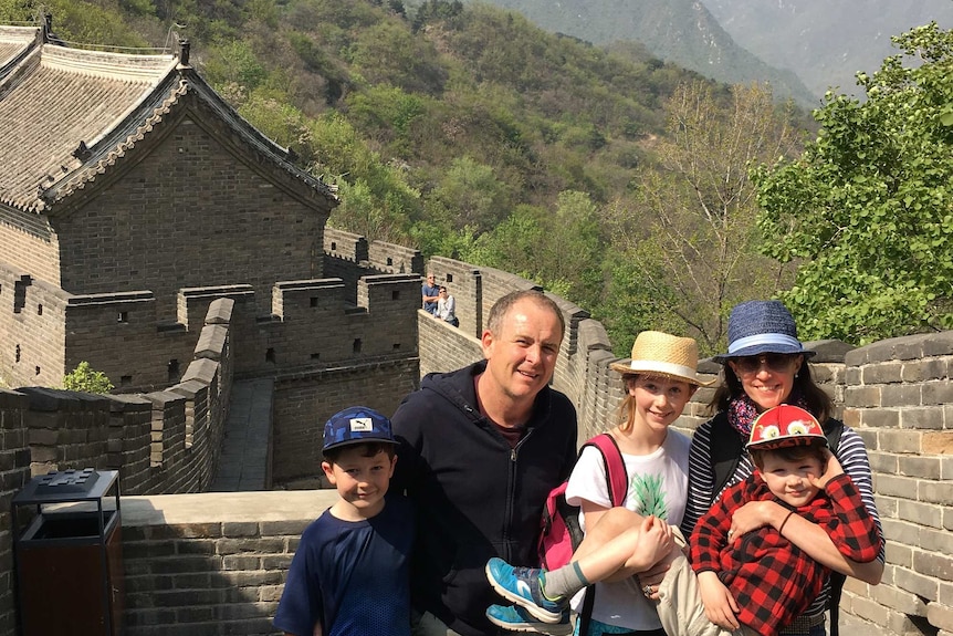 A man with his family stands on a brick wall with mountains in the background