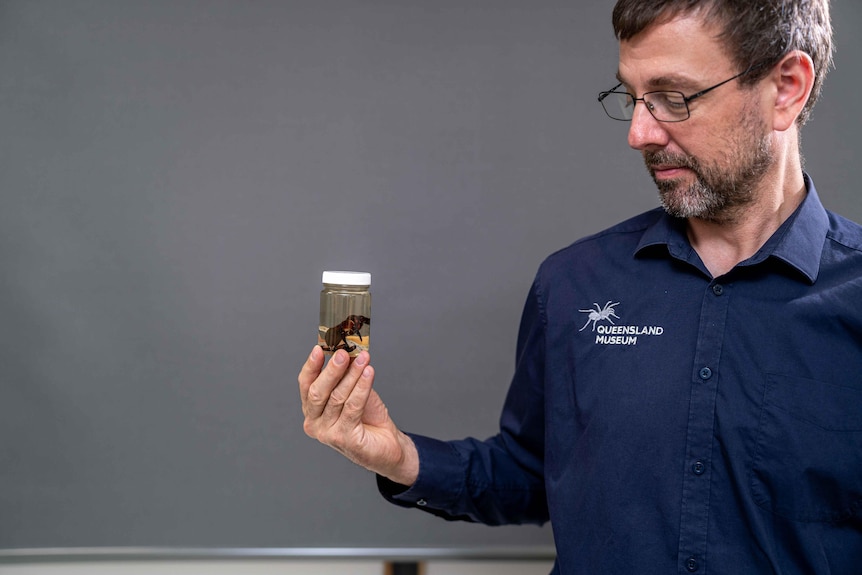 A bespectacled man with dark hair and a salt-and-pepper beard looks at a specimen in a vial in his hand.