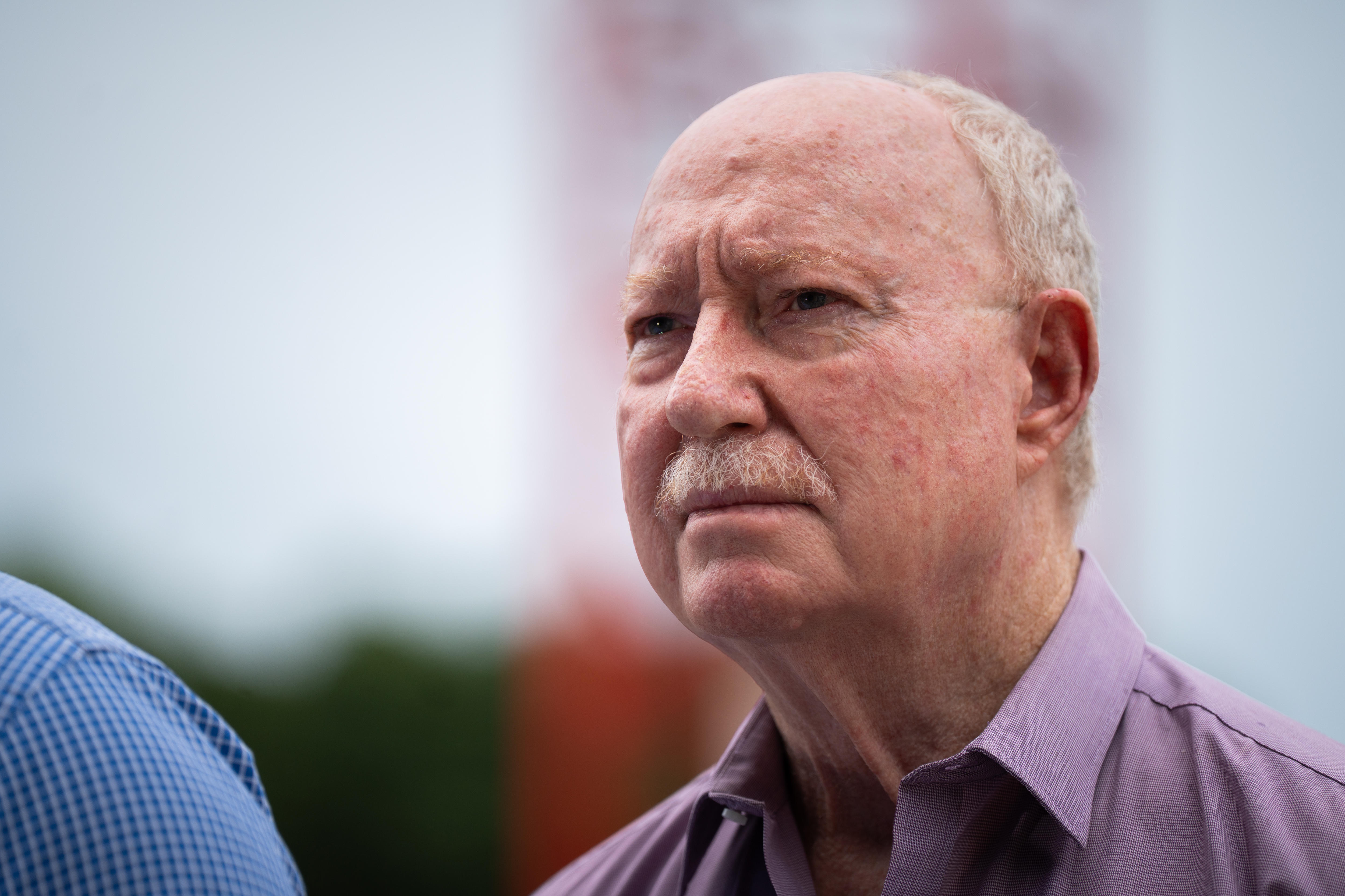 A man with grey hair and a moustache wears a serious facial expression.