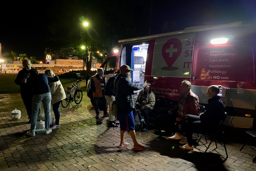 A group of people sit and stand outside a van at night.