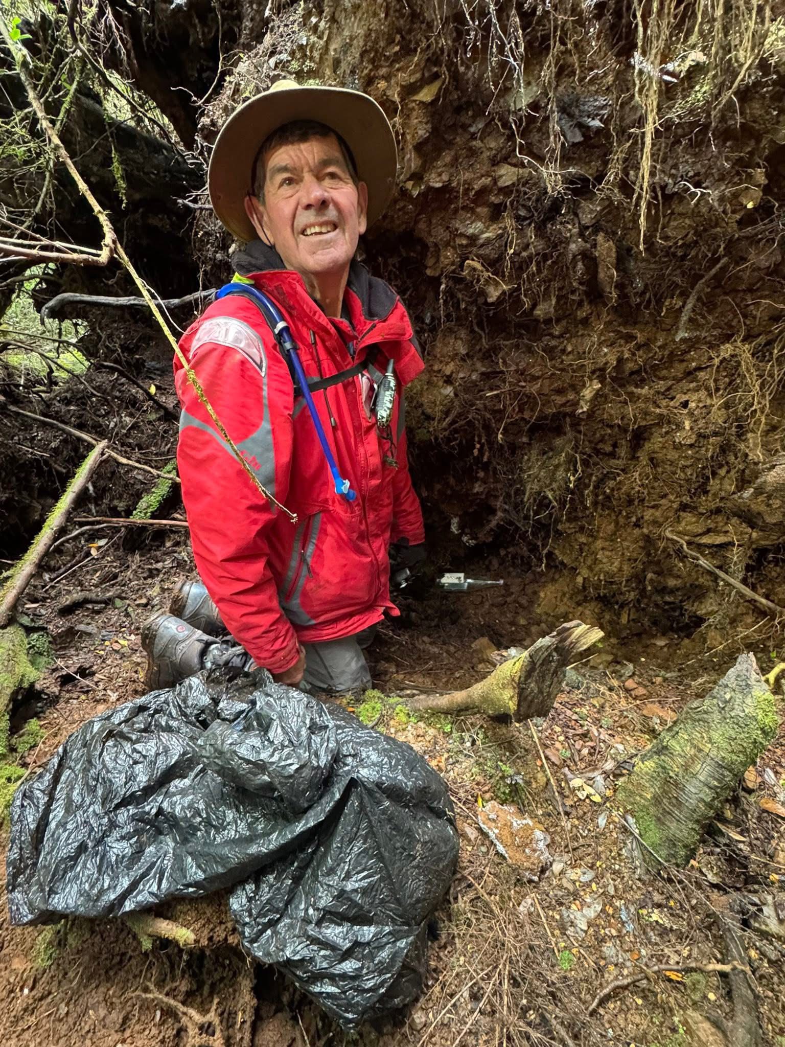 Un hombre se arrodilla junto a las raíces de los árboles, una botella de vidrio y una bolsa de basura.