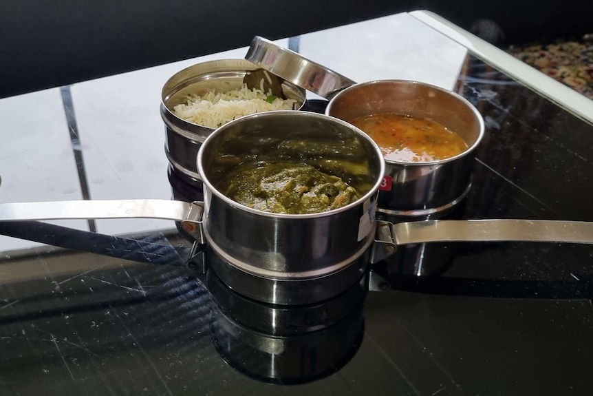An Indian-style stainless steel lunch box with three bowls stacked on a shiny table, filled with food.