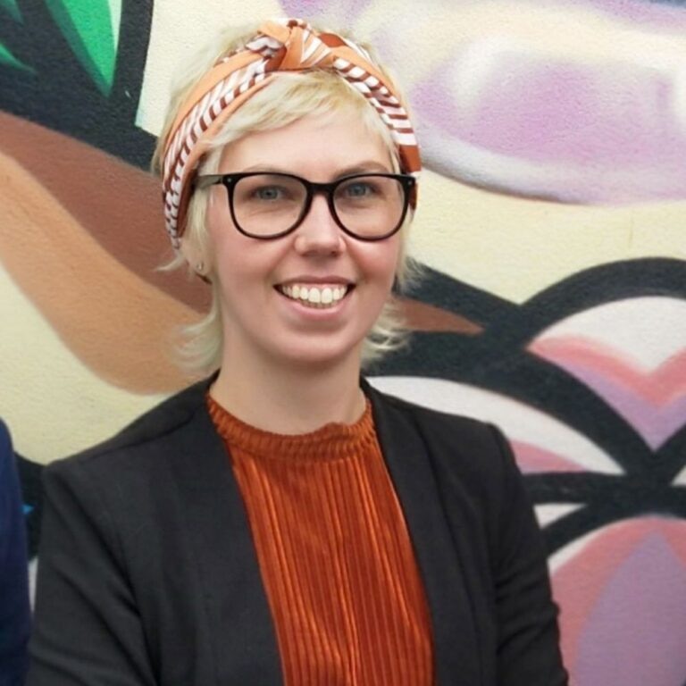 A woman smiling wearing an orange top, black glasses and a colourful scarf