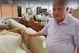 An older man gestures towards a baroque-style armchair covered in transparent plastic, furniture in background.