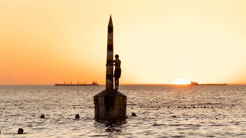 A person in silhouette stands on the pylon as the sun sets over at Cottesloe Beach on a hot day.