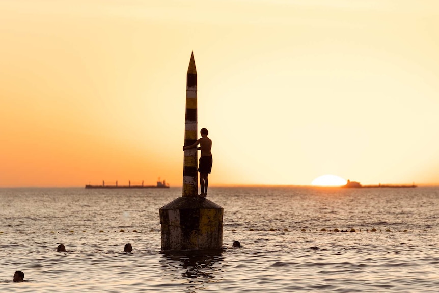 A person in silhouette stands on the pylon as the sun sets over at Cottesloe Beach on a hot day.