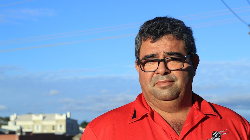 A middle-aged dark haired man in glasses and a red shirt, with blue sky behind him.