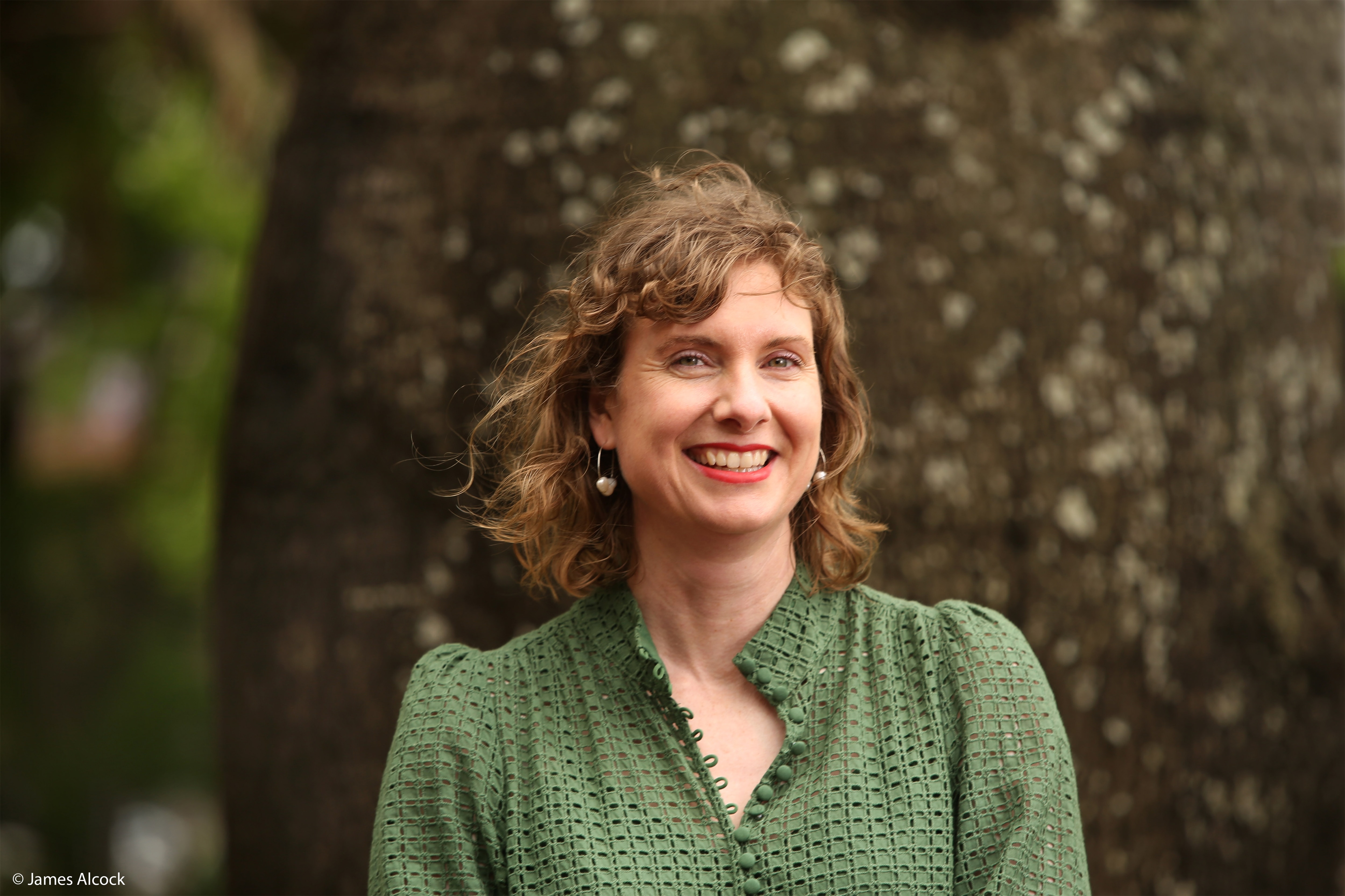 A escritora Clare Fletcher sorri, parada em frente a uma árvore, vestindo uma blusa verde e cabelo castanho curto.
