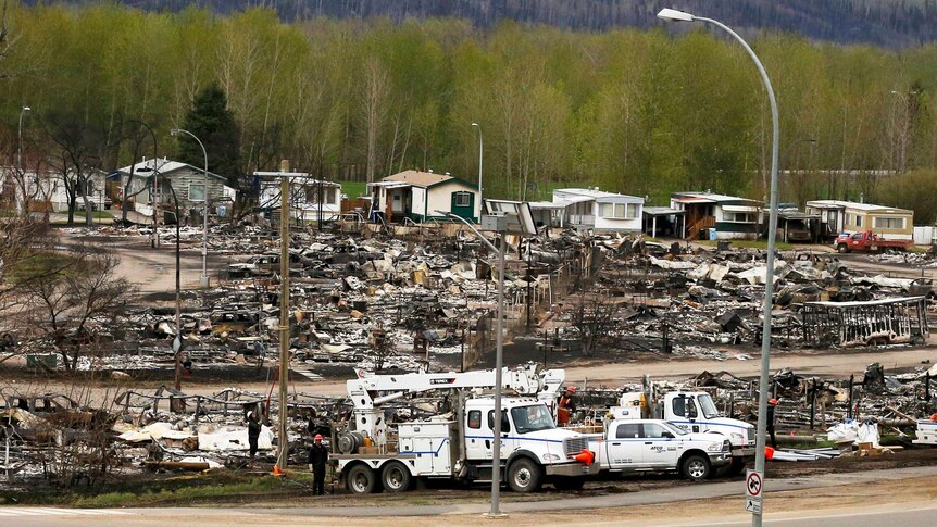 Emergency vehicles parked in front of a row of houses burned to the ground, with houses standing behind it.